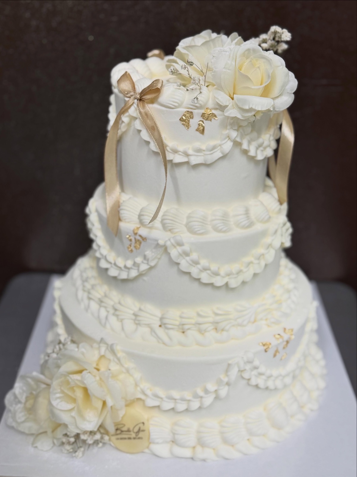 Torta nuziale a quattro piani, panna bianca lavorata a balze, rose avorio, nastri di raso e foglia oro